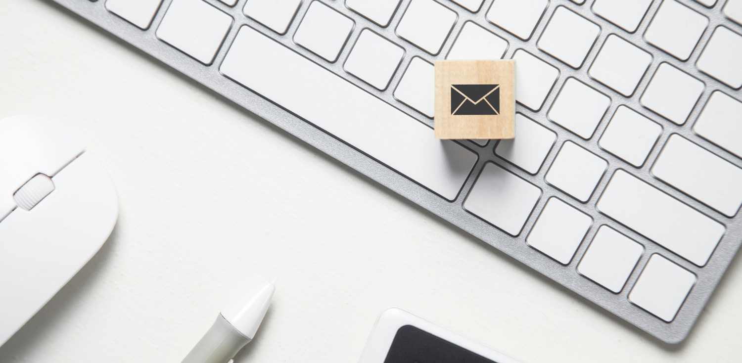 Λευκό πληκτρολόγιο, λευκό ποντίκι υπολογιστή, λευκό στυλό, μεένα τατράγωνο ξύλινο κύβο με το σύμβολο του email marketing για ένα ιστότοπο που έχει περιεχόμενο για το κόστος του email marketing.