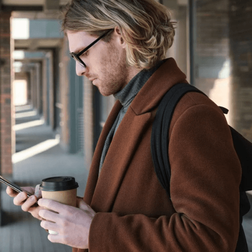 A man with blonde hair and glasses stands outdoors, looking at his phone while holding a cup of coffee. He is wearing a brown coat and has a backpack, appearing focused as he navigates a corridor of modern buildings