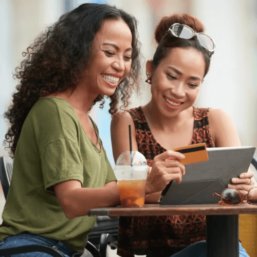 Two smiling women sitting at an outdoor table, sharing a tablet while one of them holds a credit card. A cold drink with a straw is also on the table, indicating they are likely enjoying a casual moment while shopping or browsing online