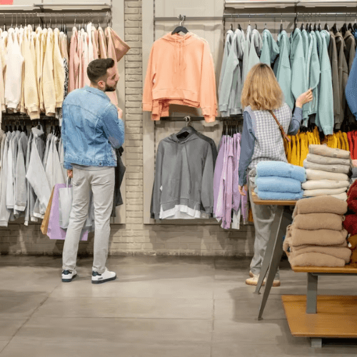 Two shoppers, a man and a woman, browsing clothing in a retail store. They are looking at colorful hoodies and sweatshirts on display racks. Folded clothes are neatly stacked on a table in the foreground, creating a cozy shopping atmosphere