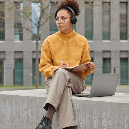 A young woman wearing glasses and headphones sits outside on a concrete ledge, holding a notebook and pen, with a laptop beside her. She is dressed in a cozy yellow sweater and light brown pants, appearing focused as she works in a peaceful urban environment.