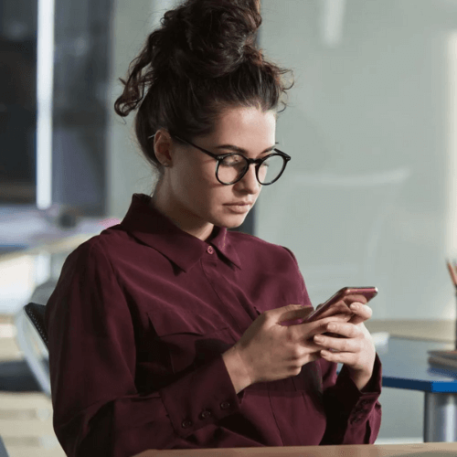 A young woman with dark hair in a bun, wearing glasses and a burgundy shirt, is focused on her phone while sitting in a well-lit office space. She appears engaged with her device, with a soft, professional environment in the background